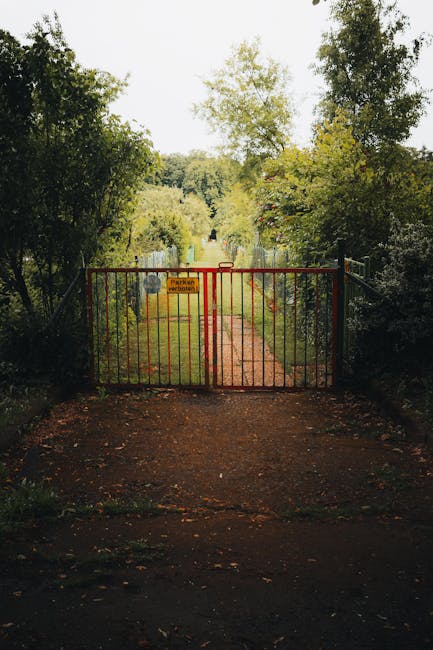 A red metal gate blocks a narrow grass pathway leading into a lush, green garden area, with tall trees and dense foliage on either side. The pathway is bordered by overgrown plants and shrubs, with so