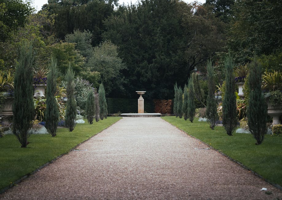 A red metal gate blocks a narrow grass pathway leading into a lush, green garden area, with tall trees and dense foliage on either side. The pathway is bordered by overgrown plants and shrubs, with so